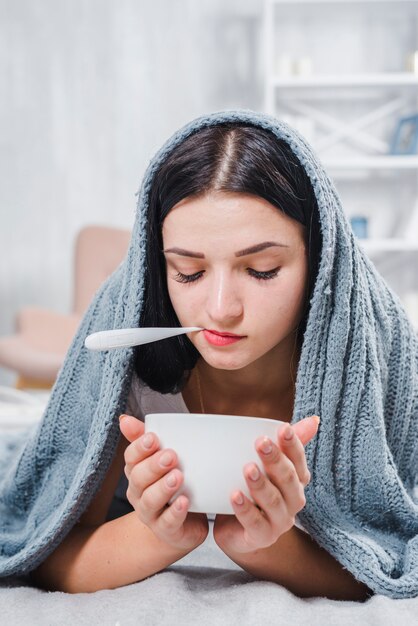 Jolie jeune femme avec foulard autour de la tête tenant un bol avec thermomètre numérique dans sa bouche