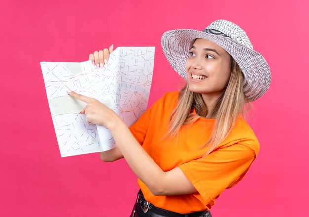 Une jolie jeune femme heureuse dans un t-shirt orange portant un chapeau pointant sur une carte avec l'index sur un mur rose