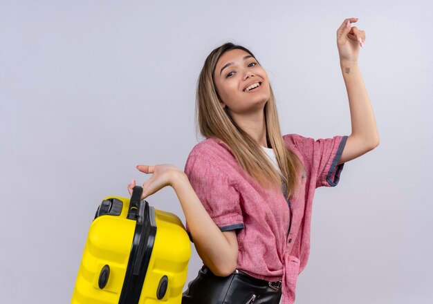 Une jolie jeune femme heureuse portant une chemise rouge tenant une valise jaune tout en regardant sur un mur blanc