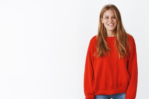 Jolie jeune femme heureuse et sympathique avec de jolies taches de rousseur et une longue coiffure en pull rouge chaud souriant largement, posant détendue et insouciante avec une expression douce et extravertie sur un mur blanc