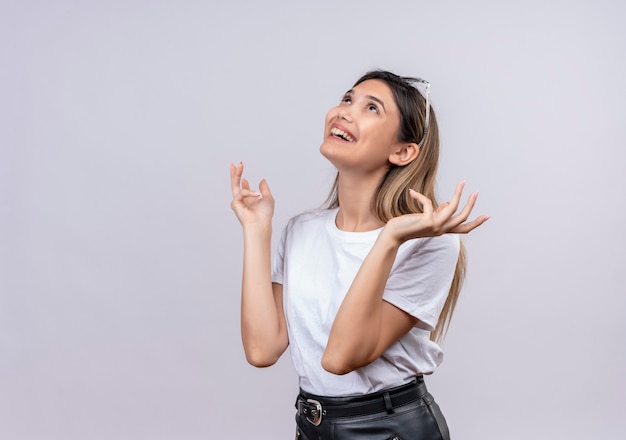 Une jolie jeune femme heureuse en t-shirt blanc portant des lunettes de soleil sur sa tête en pensant et en levant les mains dans le bonheur tout en regardant sur un mur blanc