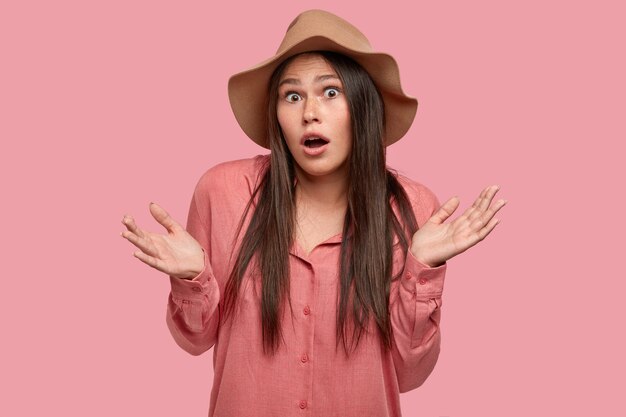 Jolie jeune femme insouciante avec de longs cheveux raides et foncés, a une expression inconsciente, serre les mains avec hésitation, ouvre la bouche de surprise