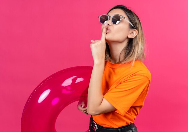 Une jolie jeune femme joyeuse dans un t-shirt orange portant des lunettes de soleil montrant le geste chut avec l'index tout en maintenant l'anneau gonflable rose sur un mur rose