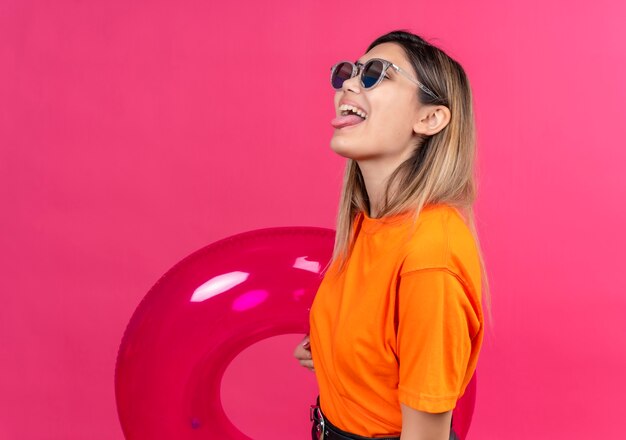 Une jolie jeune femme joyeuse dans un t-shirt orange portant des lunettes de soleil montrant sa langue tout en tenant un anneau gonflable rose sur un mur rose