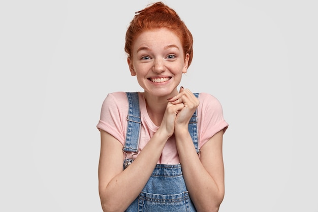 Jolie jeune femme joyeuse à tête rouge avec une expression faciale joyeuse, garde les mains jointes, a les yeux bleus pleins de bonheur, se tient contre un mur blanc, reçoit un joli cadeau