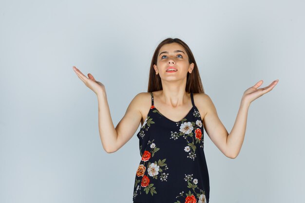 Jolie jeune femme levant les paumes écartées tout en levant les yeux en chemisier et en ayant l'air reconnaissante. vue de face.