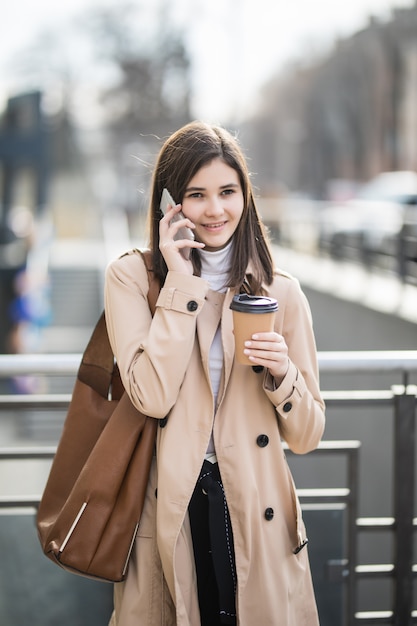 Jolie jeune femme marchant dans la rue en tenant la tasse de café et le téléphone