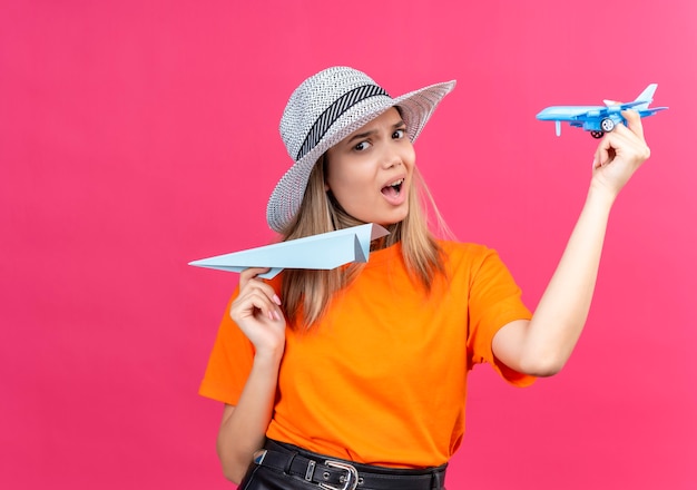 Photo gratuite une jolie jeune femme mécontente dans un t-shirt orange portant un chapeau volant avion en papier tout en tenant un avion jouet bleu sur un mur rose