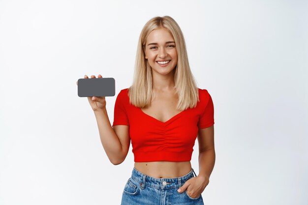 Jolie jeune femme montrant un écran horizontal de téléphone portable et souriant recommandant un site Web ou une application de shopping fond blanc