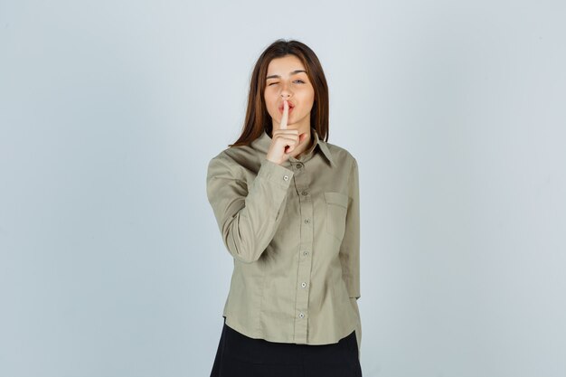 Jolie jeune femme montrant un geste de silence tout en clignant des yeux en chemise, jupe et semblant raisonnable. vue de face.