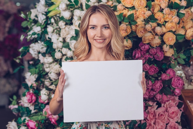 Jolie jeune femme montrant une pancarte blanche blanche dans la main debout contre des roses colorées