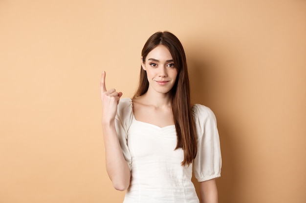 Jolie jeune femme montre le doigt numéro un souriant et l'air confiant debout sur fond beige...