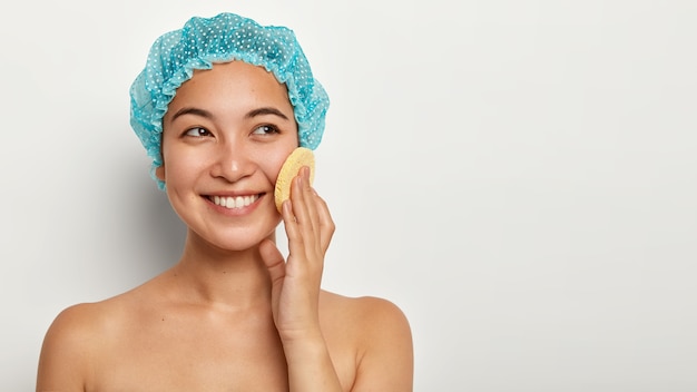 Jolie jeune femme nettoie le visage avec une éponge, touche la joue, regarde de côté porte un bonnet de bain, a une apparence agréable