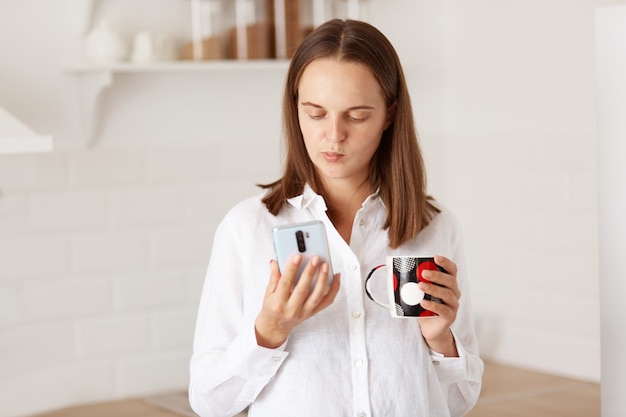 Jolie jeune femme pensive adulte debout avec un téléphone intelligent dans les mains, lisant des nouvelles sur les réseaux sociaux, buvant du café ou du thé dans la cuisine, vêtue d'une chemise blanche de style décontracté.