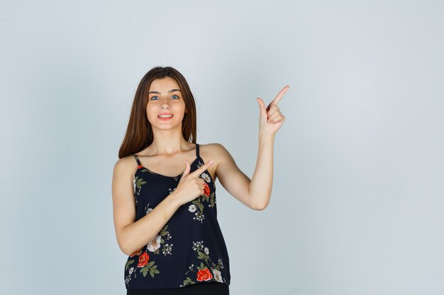 Jolie jeune femme pointant vers le coin supérieur droit en blouse et l'air heureux. vue de face.