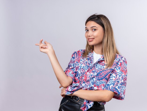 Une jolie jeune femme portant une chemise imprimée paisley montrant quelque chose tout en regardant sur un mur blanc