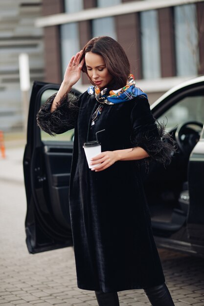 Jolie jeune femme portant un manteau noir à la mode tout en gardant le café et debout près de la voiture. Concept de ville de mode