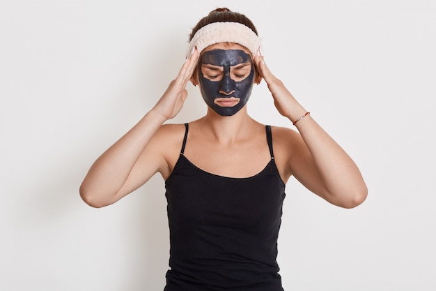 Jolie jeune femme posant les yeux fermés et gardant les mains sur les tempes, souffrant de douleur, portant un bandeau et un t-shirt sans manches, faisant des procédures cosmétiques à la maison, posant avec un masque de boue.