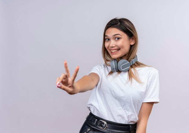 Une jolie jeune femme positive en t-shirt blanc dans les écouteurs montrant le geste de deux doigts tout en regardant sur un mur blanc