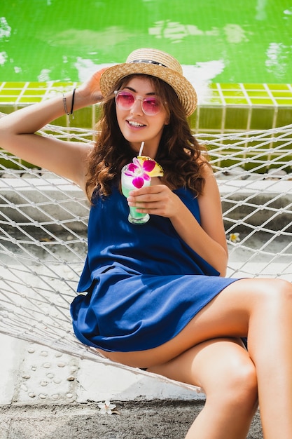 Jolie Jeune Femme En Robe Bleue Et Chapeau De Paille Portant Des Lunettes De Soleil Roses, Boire Un Cocktail D'alcool En Vacances Assis Dans Un Hamac En Tenue De Style Estivale