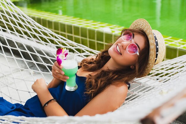 Jolie jeune femme en robe bleue et chapeau de paille portant des lunettes de soleil roses, boire un cocktail d'alcool en vacances assis dans un hamac en tenue de style estivale