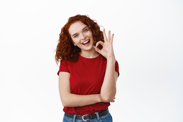 Jolie jeune femme rousse aux cheveux bouclés, tête inclinée mignonne, montrant un signe OK et souriante heureuse, approuvant et comme une bonne chose, qualité de louange, debout sur blanc