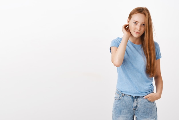 Jolie jeune femme rousse européenne avec des taches de rousseur effleurant les cheveux derrière l'oreille tenant la main dans la poche et regardant avec une expression timide et maladroite