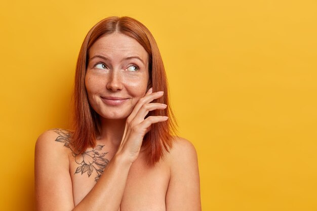 Jolie jeune femme rousse touche doucement la joue, regarde avec une expression rêveuse et réfléchie de côté, se tient nue, a un corps bien soigné et une peau saine, des modèles contre le mur jaune, un espace vide