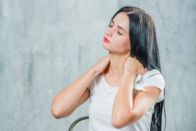 Jolie jeune femme souffrant de douleurs au cou, assise contre un mur gris