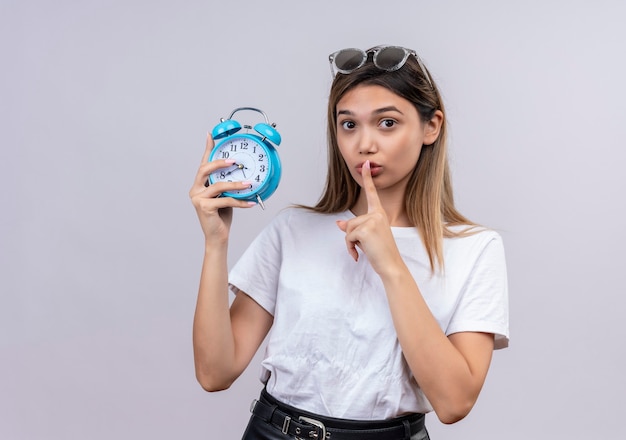 Une jolie jeune femme en t-shirt blanc à lunettes de soleil montrant le geste chut tout en tenant un réveil bleu sur un mur blanc