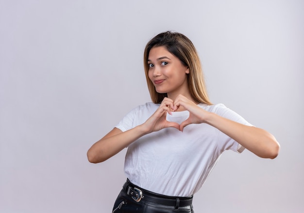 Une jolie jeune femme en t-shirt blanc montrant le signe en forme de coeur exprimant le symbole du mariage et de la santé sur un mur blanc