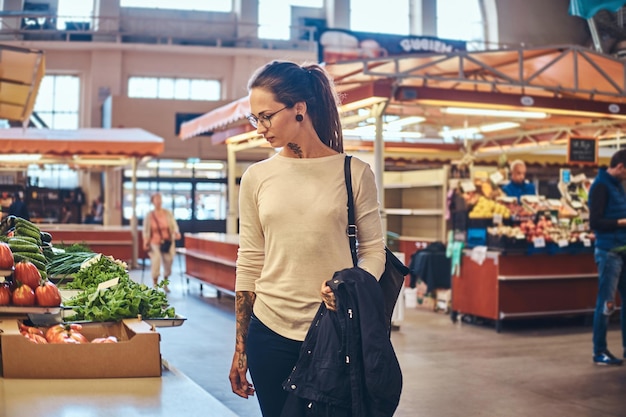 Jolie jeune femme avec tatouage et lunettes visitant le marché local pour les légumes frais.