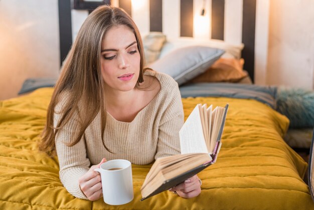 Jolie jeune femme tenant une tasse de café dans le livre de lecture à la main