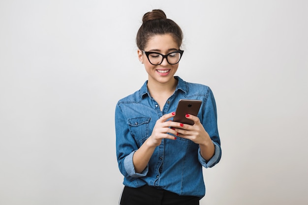 Jolie jeune femme tenant un téléphone intelligent, à l'aide de l'appareil, portant des lunettes élégantes, souriant, isolé, chemise en jean