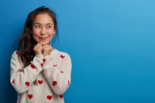 Jolie jeune femme voit quelque chose de merveilleux, tentant d'acheter, garde les mains sous le menton, regarde avec le sourire de côté, se souvient d'un moment agréable, habillé en pull décontracté, pose contre le mur bleu