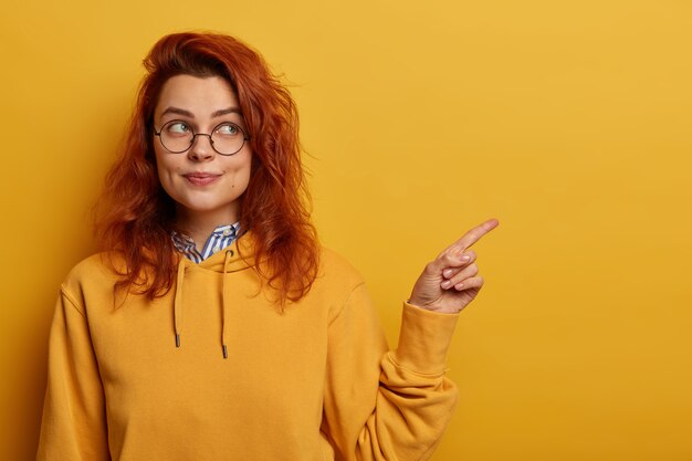 Jolie jeune fille au gingembre pointe l'index sur la gauche à l'espace vide, annonce quelque chose, porte des lunettes optiques et un sweat-shirt jaune