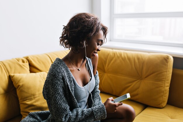 Jolie jeune fille aux cheveux bouclés courts écouter de la musique dans des écouteurs, avec un téléphone à la main, assis sur un canapé jaune avec des coussins, au repos à la maison