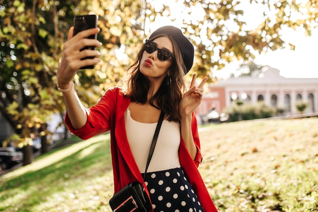 Jolie jeune fille aux cheveux bruns et aux lèvres rouges, en béret, lunettes de soleil noires, haut et chemise élégants, faisant du selfie à l'extérieur