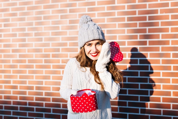 Jolie jeune fille aux cheveux longs en bonnet tricoté et gants sur le mur extérieur. Elle tient un cadeau ouvert dans ses mains, souriant.