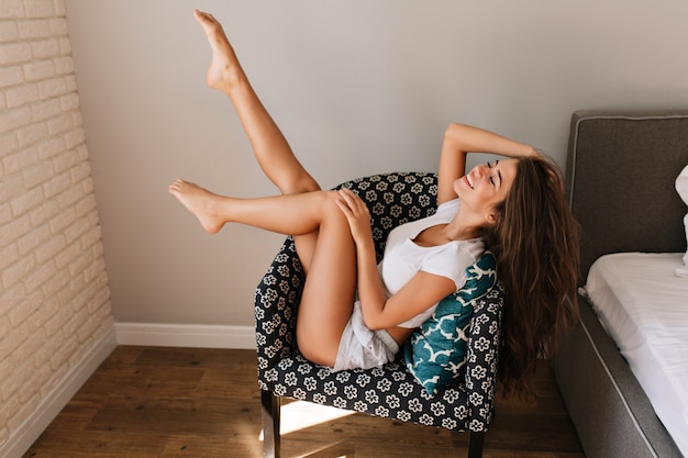 Jolie jeune fille aux cheveux longs en chemises de refroidissement dans une chaise. Elle tient les jambes au-dessus, souriant les yeux fermés.