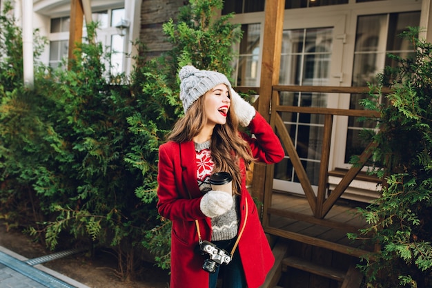 Jolie jeune fille aux cheveux longs en manteau rouge et bonnet tricoté sur maison en bois. Elle tient le café pour aller dans des gants blancs, exprimant à côté.