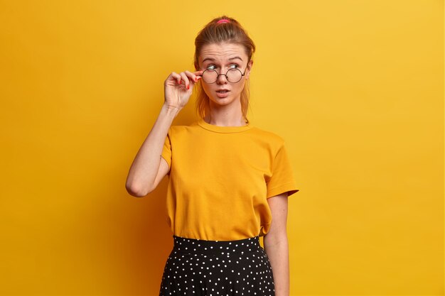 Jolie jeune fille entend des rumeurs étonnantes, garde la main sur le bord des lunettes, porte un t-shirt jaune, une jupe à pois, détourne le regard avec surprise et suspicion