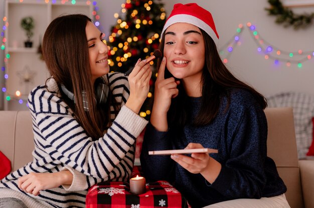 Une jolie jeune fille ravie maquille son amie avec un bonnet de noel assis sur un fauteuil et profite de la période de Noël à la maison
