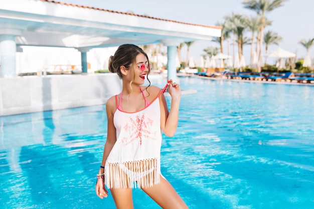 Jolie jeune fille en robe de plage debout devant la piscine extérieure avec des palmiers sur fond et en détournant les yeux