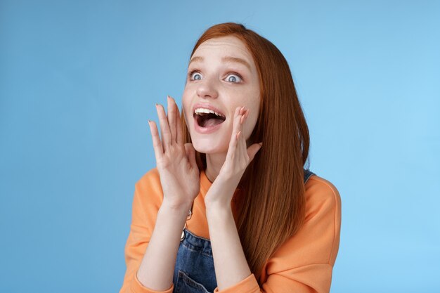 Jolie jeune fille rousse européenne stupide de 20 ans appelant un ami à la recherche de quelqu'un la foule a l'air détendu en criant joyeusement tenir les mains ouvertes la bouche criant le nom plus fort regarde à gauche, fond bleu.