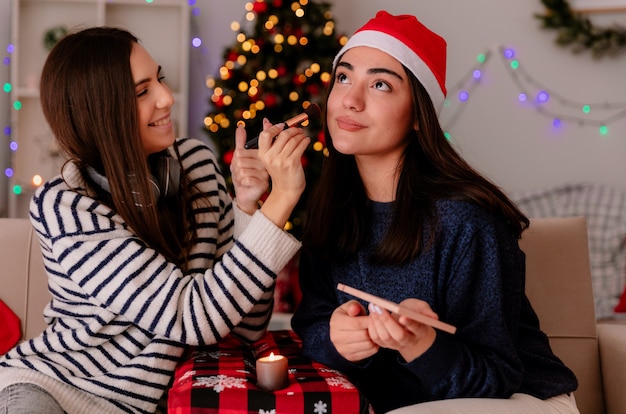 Une jolie jeune fille souriante maquille son amie heureuse avec un bonnet de noel assis sur un fauteuil et profitant de la période de Noël à la maison