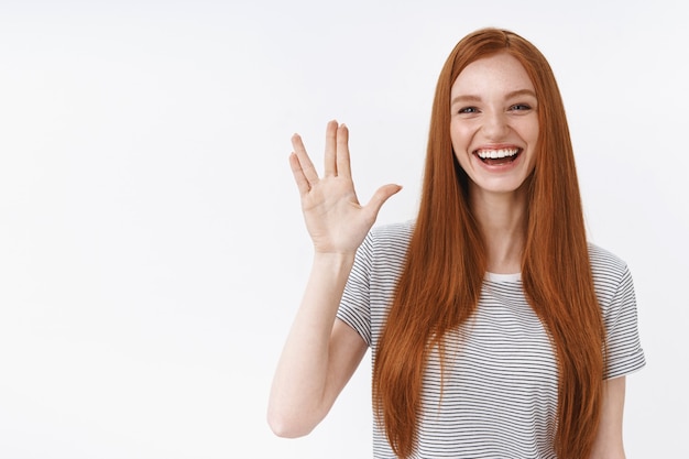 Photo gratuite jolie jeune geek rousse insouciante comme regarder des films fantastiques de fans de séries télévisées salue des amis levant le geste de la main montrant un geste souriant largement s'amuser en accueillant une fête d'invités, mur blanc