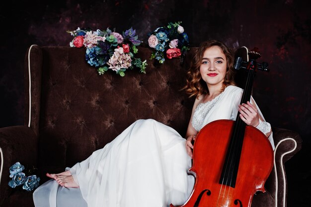 Jolie jeune musicienne gilrl en robe blanche avec contrebasse assise sur un canapé vintage marron