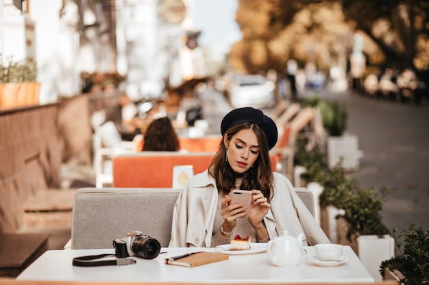 Jolie jeune photographe avec une coiffure brune ondulée, un béret, un trench-coat beige assis à la terrasse du café de la ville, prenant un thé et un gâteau au fromage, tenant et regardant dans un téléphone portable