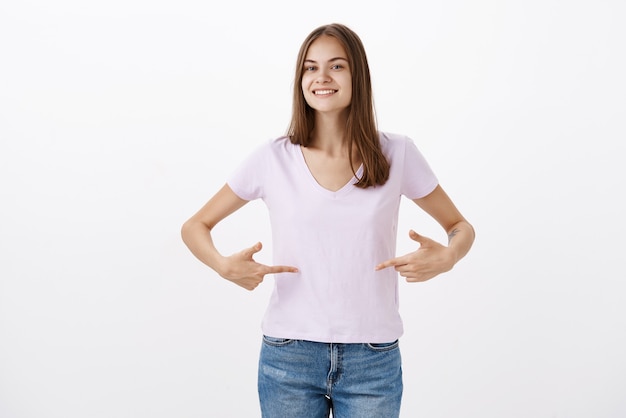 Jolie jeune sportive donnant des conseils sur la façon de rester en forme en souriant joyeusement en regardant amicalement pointant sur le t-shirt ou le ventre debout ravi et heureux avec un regard heureux sur un mur gris